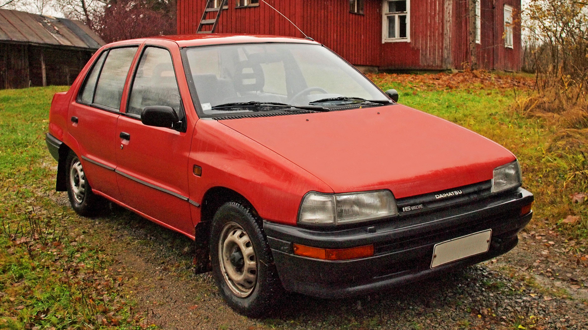 A red compact hatchback car parked on a grassy driveway, with an old wooden house and trees in the background on an overcast day.