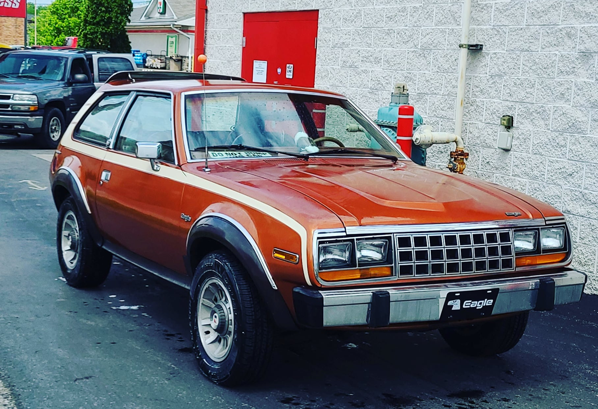 A vintage brown AMC Eagle two-door car is parked on a paved lot near a light-colored brick building, with another vehicle, a fire hydrant, and red door in the background.