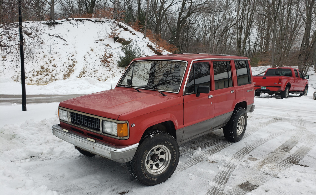 A red, boxy SUV is parked on a snowy road with trees and a snow-covered slope in the background. Another red vehicle is parked further back.