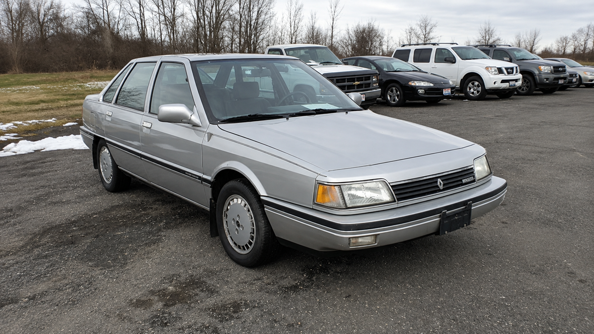 A silver four-door sedan parked in a lot with several other vehicles and bare trees in the background. The ground is partly covered with patches of snow.