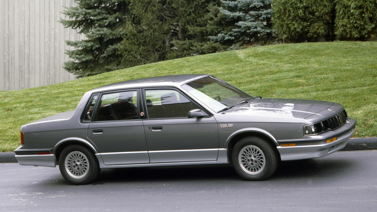 A silver four-door sedan from the 1980s is parked on a paved road with a grassy hill and evergreen trees in the background.