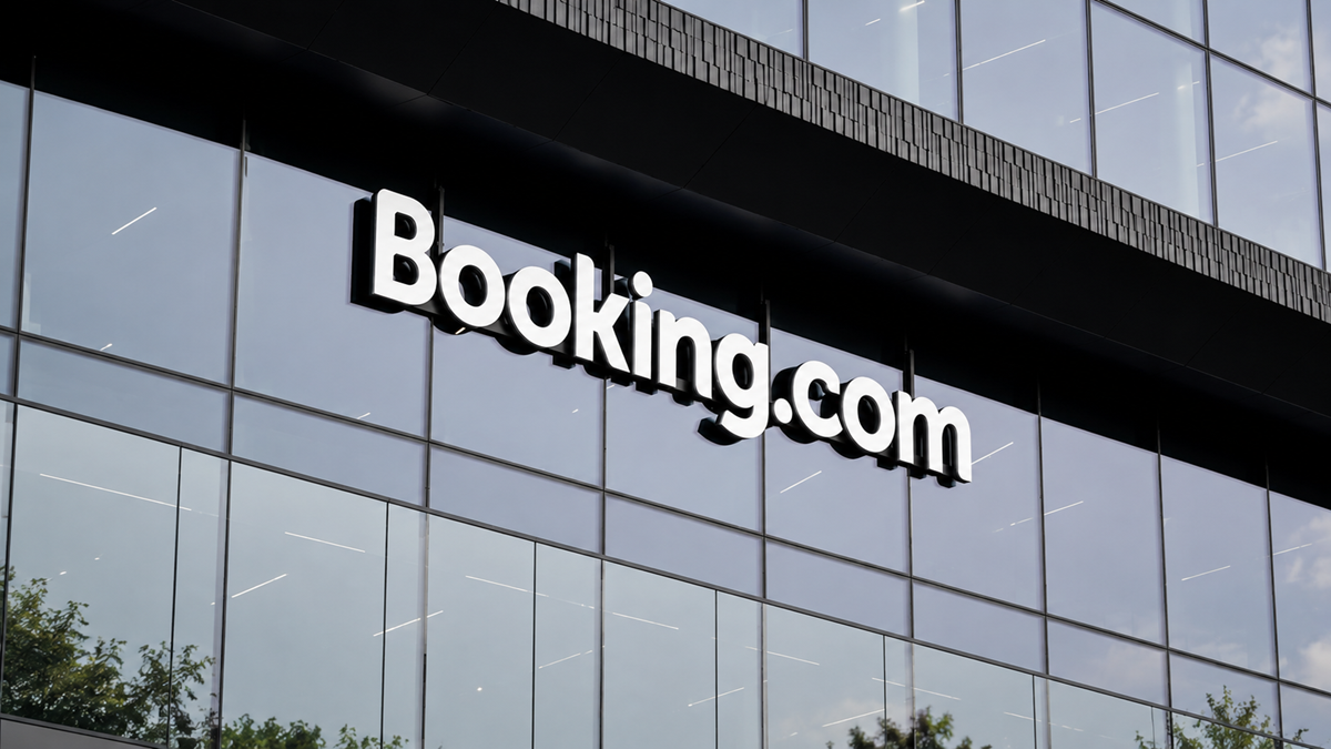 Large white “Booking.com” sign on the glass facade of a modern building, reflecting blue sky and tree branches.