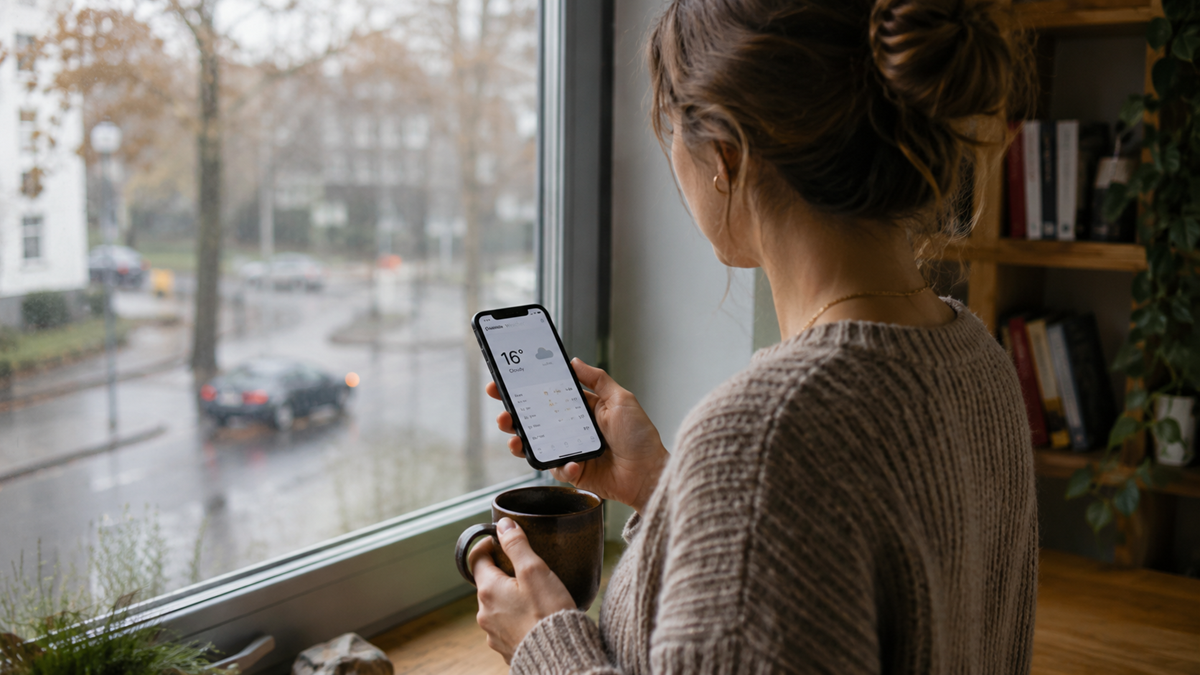 A woman stands by a window holding a mug and looking at her smartphone, which displays a weather app showing 16°C. It is rainy outside, and bookshelves are visible inside.