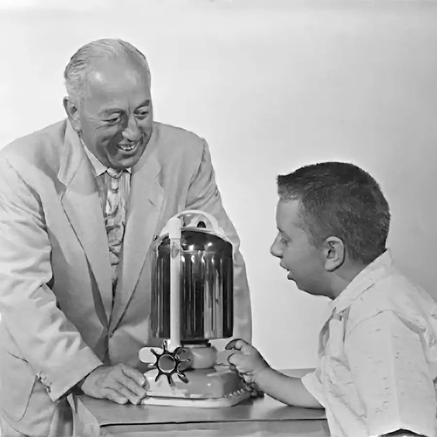 An older man and a young boy smile while looking at a shiny, vintage coffee percolator on a small table. The man wears a light suit, and the boy wears a short-sleeve shirt. The background is plain white.