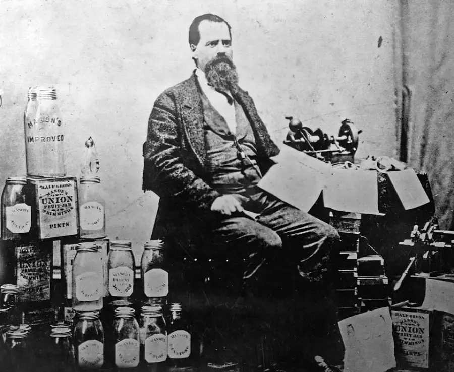 A bearded man in a suit sits near a table with bottles, jars, and papers. Several jars are labeled “Union” and “Mason’s Improved.” The setting appears to be from the late 1800s or early 1900s.