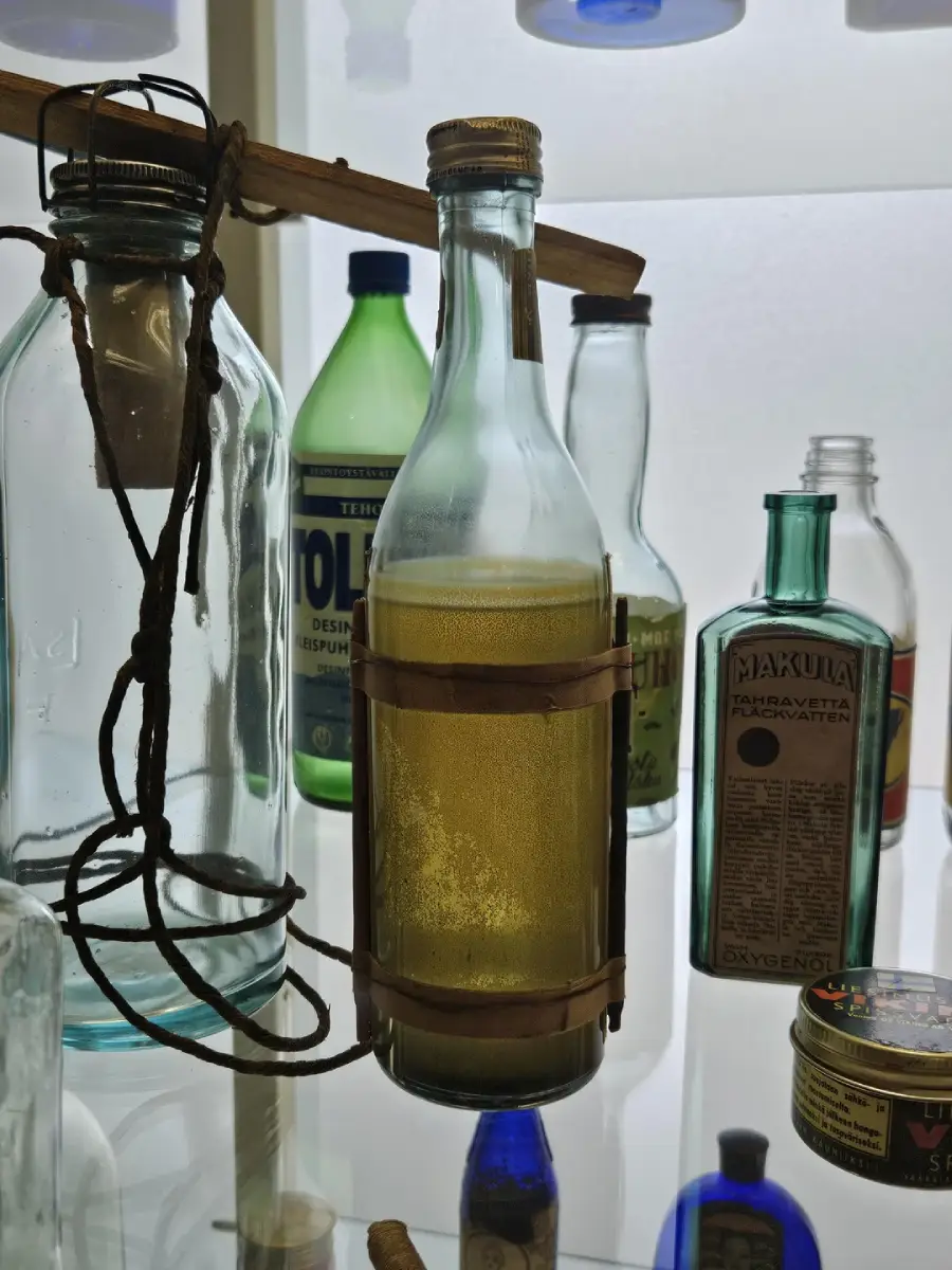 A glass bottle with brown liquid and cork cap is held by leather straps. It is displayed among various vintage bottles and tins on shelves with a white background.