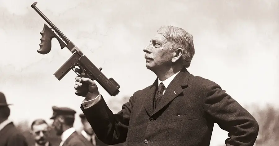 An older man in a suit holds a Thompson submachine gun pointed upwards. Several people stand in the blurred background outdoors on a clear day. The image is in black and white.