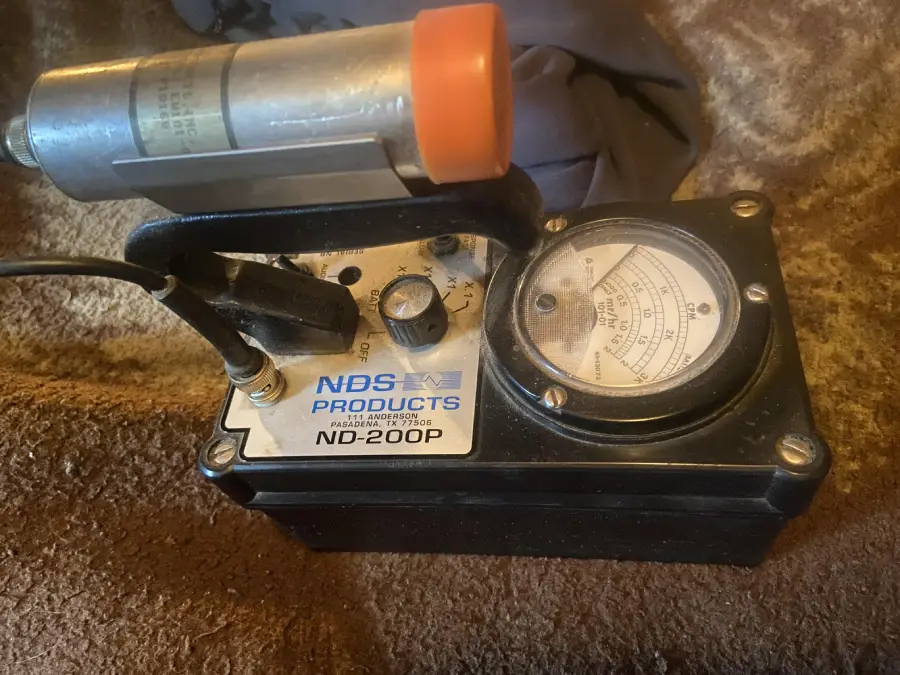 A close-up of an NBS Products ND-200P device with a dial gauge, knob, and attached cylindrical metal component, resting on a brown carpet.