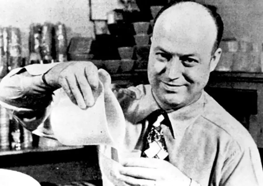 A man in a dress shirt and tie smiles while pouring liquid from a pitcher into a cup, with shelves of containers in the background.