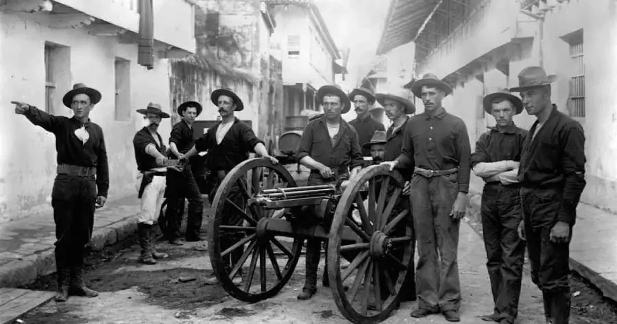A group of men in old-fashioned military uniforms stand around a Gatling gun on a street, with one man pointing to the left. The scene appears historical, with worn buildings in the background.