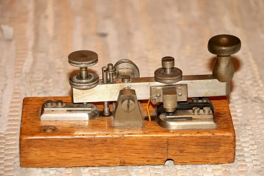 A vintage metal and wood telegraph key rests on a light patterned surface, used historically for sending Morse code messages.