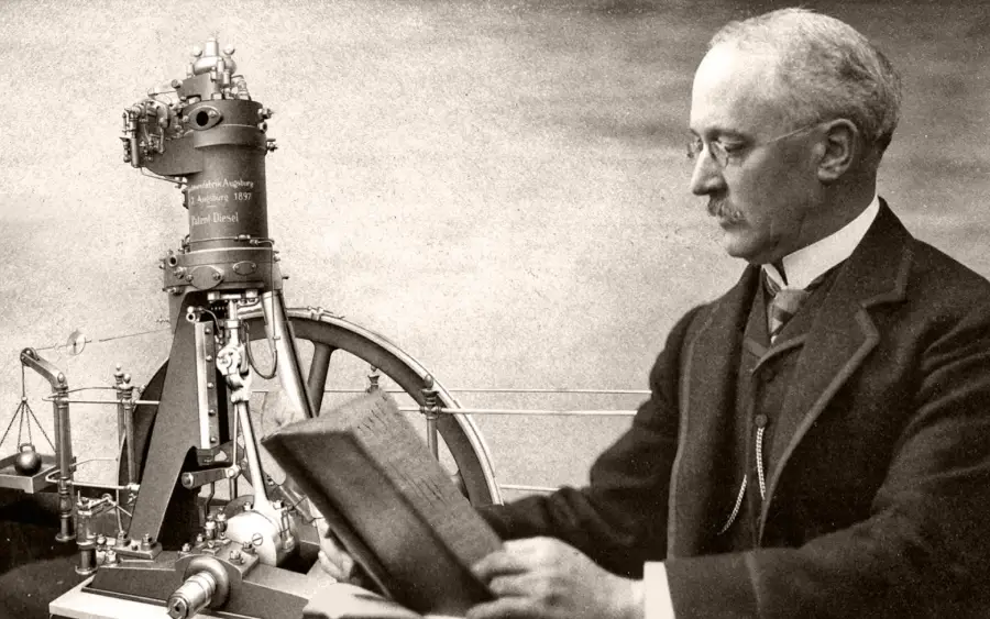 A man in a suit reads a book while sitting next to an early diesel engine model on a table, with a plain backdrop behind them.