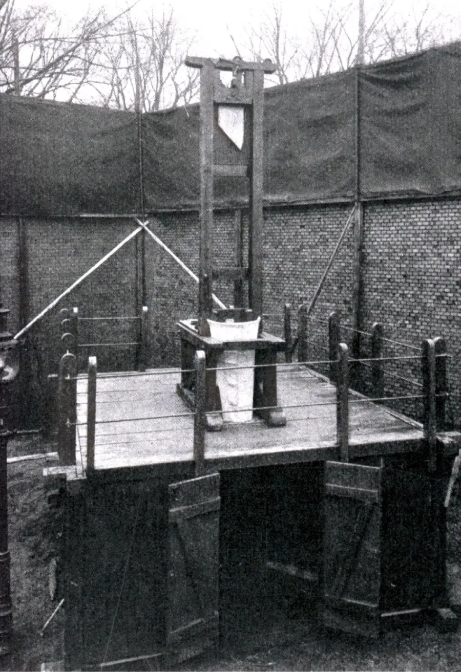 A black-and-white photo of a guillotine set on a wooden platform outdoors, surrounded by fencing and trees in the background.