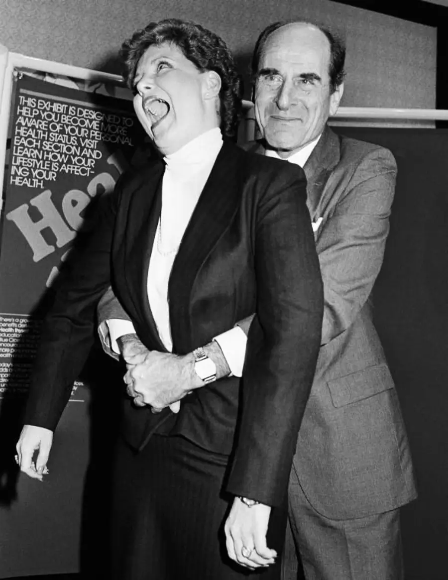 A man demonstrates the Heimlich maneuver on a smiling woman in a suit, in front of a health awareness poster. The image appears to be from a public demonstration or educational event.