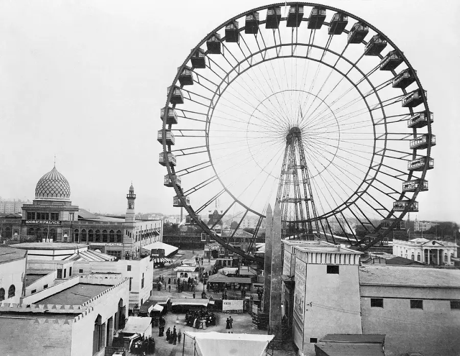 A large Ferris wheel towers over a busy fairground with people, tents, and buildings, including one with a dome and minaret, in an old black and white photograph.