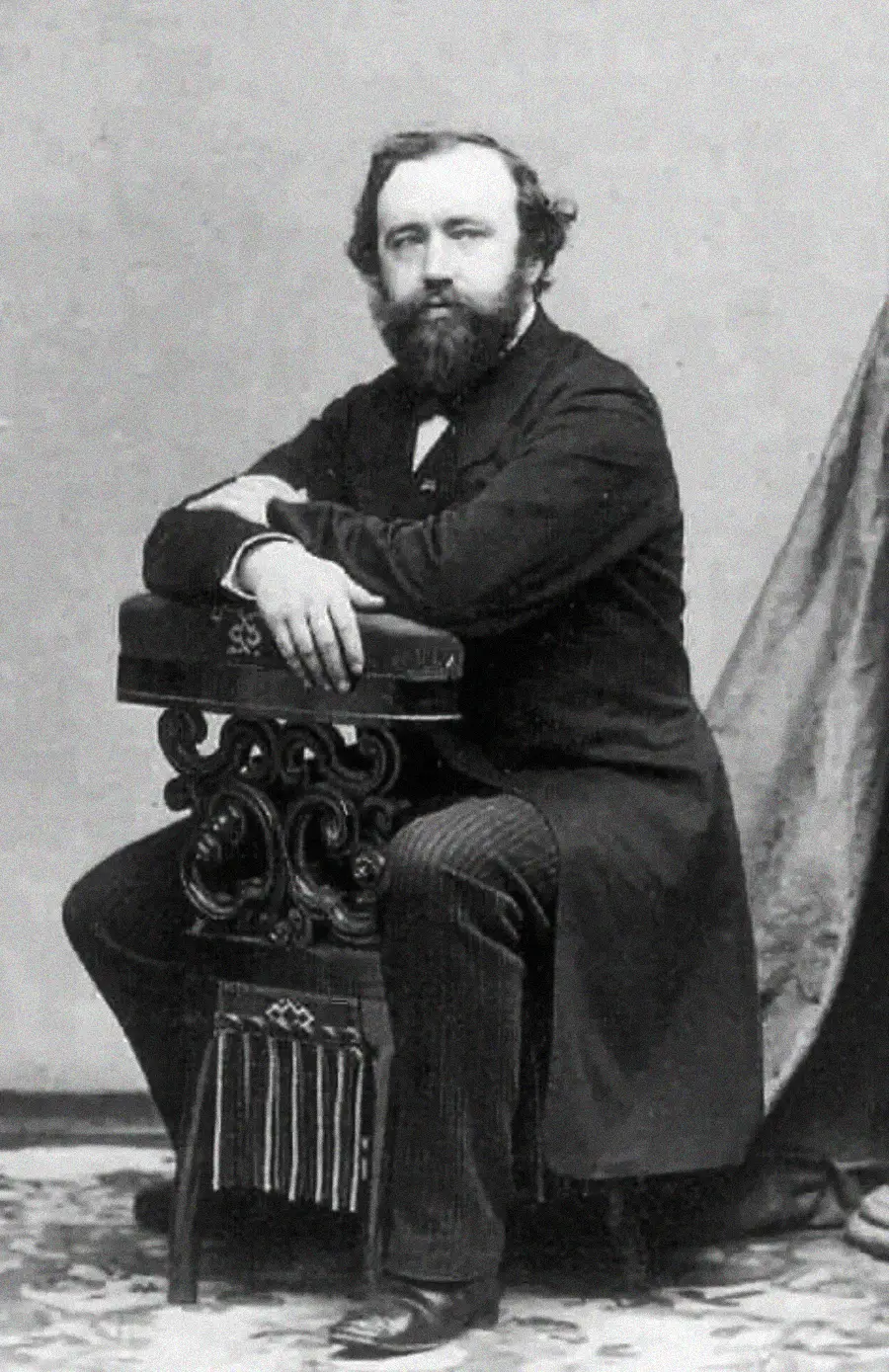A bearded man in 19th-century attire sits with one arm resting on an ornate stand, looking at the camera.
