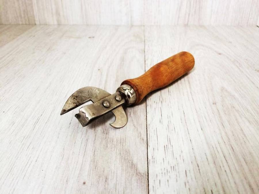 A vintage can opener with a wooden handle and metal blade is lying on a light wooden surface.