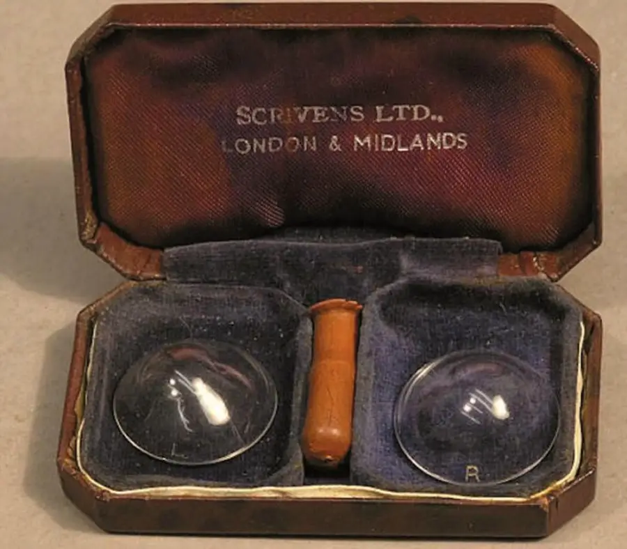 An open vintage case from Scrivens Ltd. holds two glass contact lenses and a small brown tool, all placed in a blue velvet-lined interior.