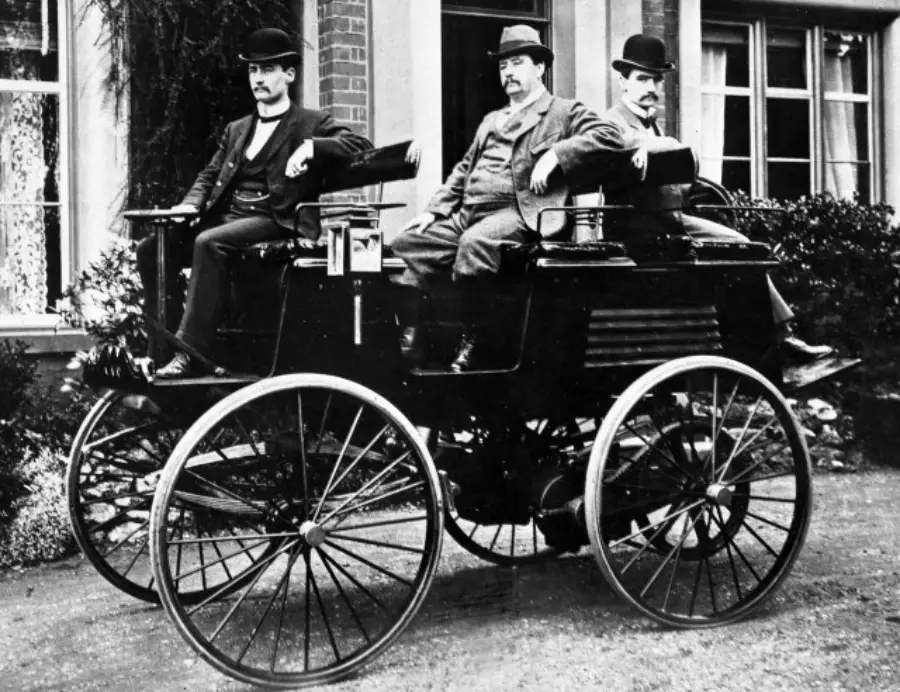 Three men in suits and bowler hats sit on an old-fashioned, open-top early automobile with large wheels, parked in front of a building with windows and shrubbery.