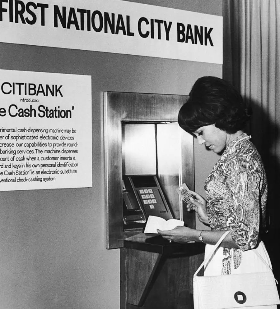A woman uses an early ATM at First National City Bank, holding cash and a receipt, next to a sign introducing the “Citibank Cash Station.” The photo is in black and white and appears to be from the 1970s.