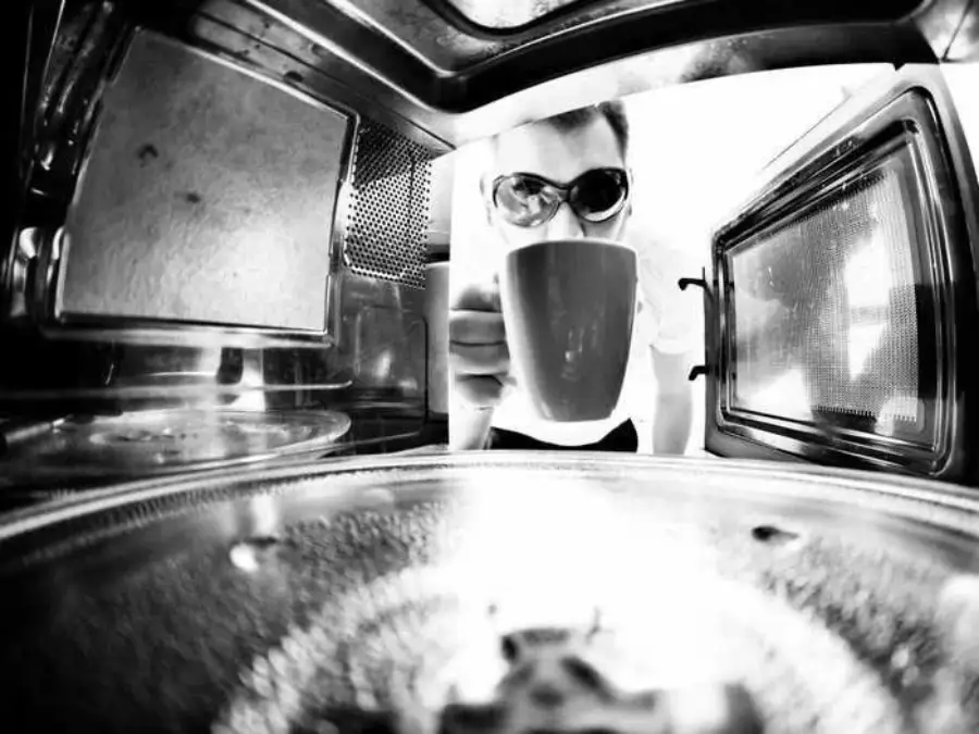 A person wearing sunglasses holds a mug, seen from inside an open microwave oven. The scene is in black and white.