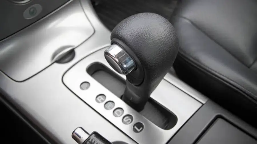 Close-up of a black automatic gear shift in a car, surrounded by a silver panel, with gear positions visible and part of a leather seat next to it.