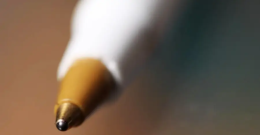 Close-up image of the tip of a ballpoint pen with a white barrel and gold-colored tip, shown against a blurred background.