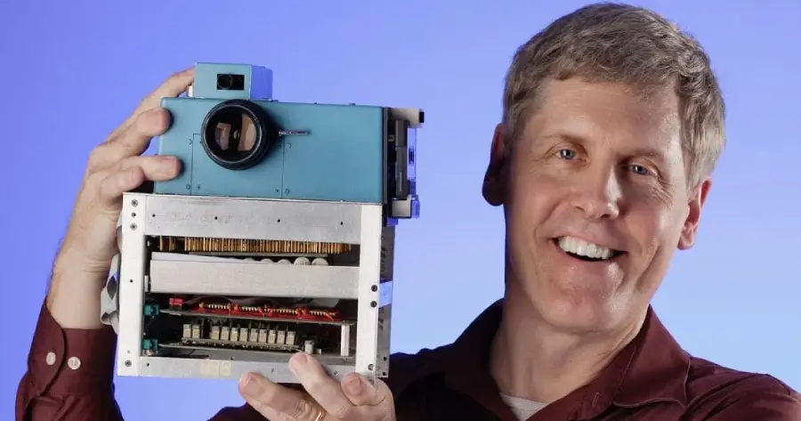 A man smiling and holding an early, bulky digital camera with visible electronic components against a blue background.