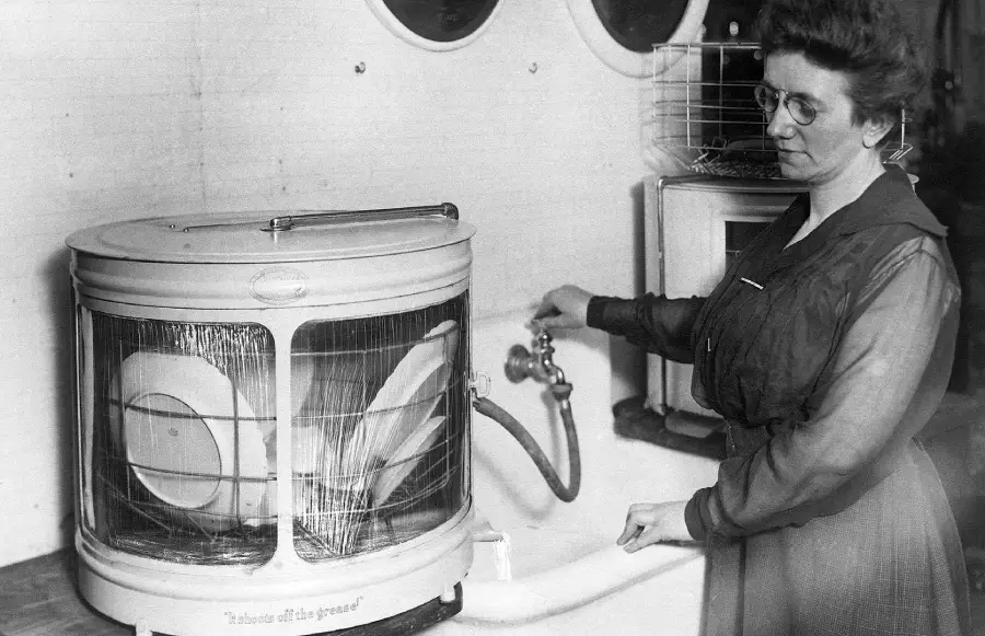 A woman in glasses stands beside an old-fashioned dishwasher filled with plates, turning a faucet to fill the machine. The dishwasher has a round, transparent door and is placed in a kitchen setting.