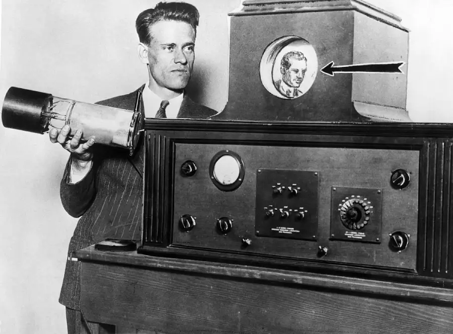 A man stands beside an early television system, holding a device and looking at a small screen displaying a person’s face, with an arrow pointing to the screen.