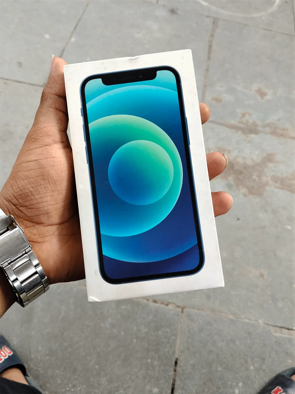 A hand holding a sealed box of a blue smartphone, likely an iPhone, with a graphic of the phone’s screen on the front. The background is gray stone pavement.