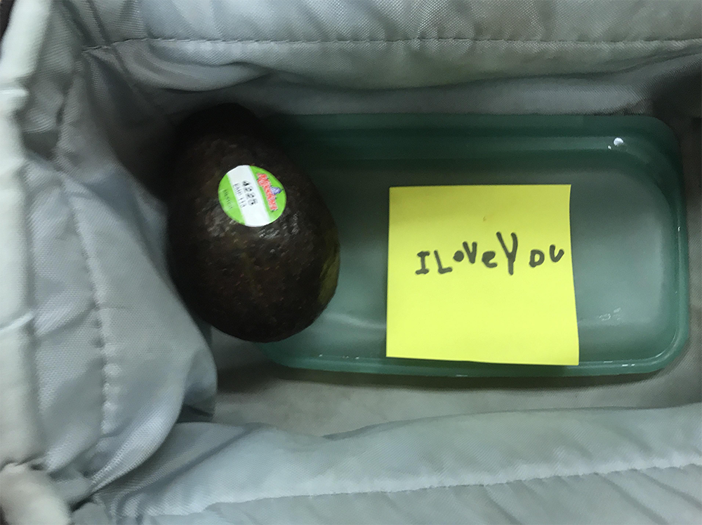 A ripe avocado and a glass container with a yellow sticky note reading “I LOVE YOU” are inside a gray lunch bag.