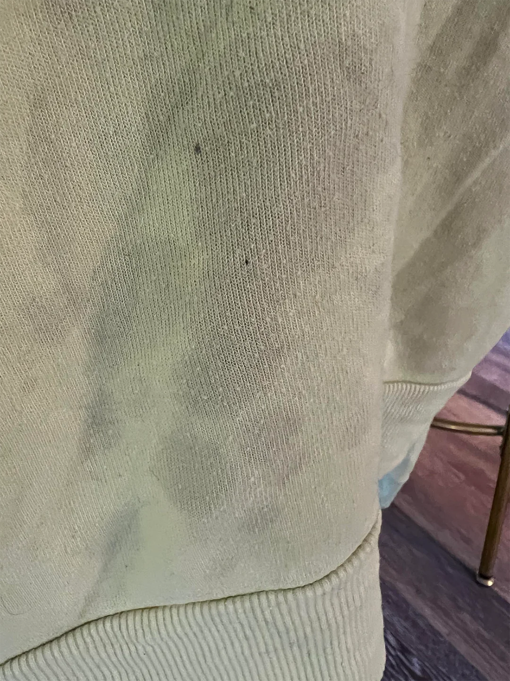 Close-up of a light-colored fabric, possibly a sweater or sweatshirt, showing subtle stains and a ribbed hem at the bottom. Part of a metal chair leg and dark wooden floor are visible in the background.