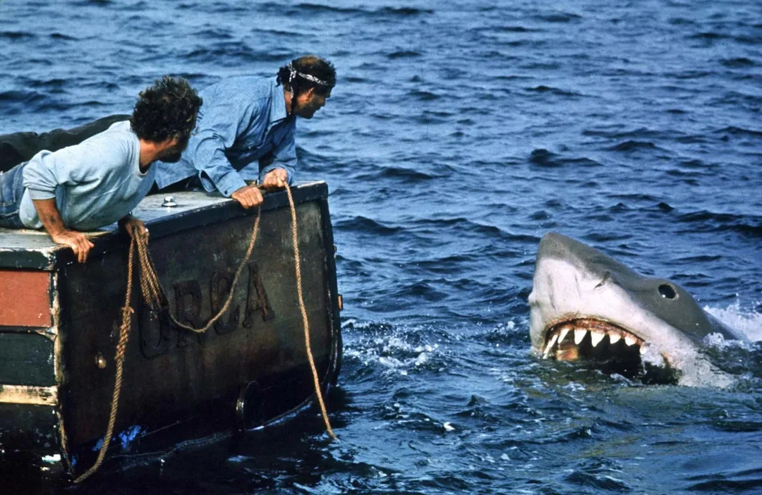 Two people on a small boat lean over the edge, staring at a large shark with its mouth open wide as it emerges from the water.