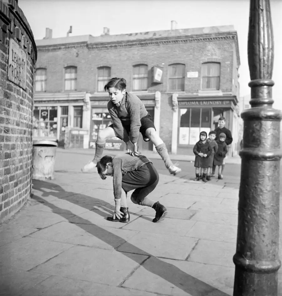 Two boys play leapfrog on a city sidewalk while a group of children watches from a distance. Old brick buildings and a streetlamp are visible in the background.