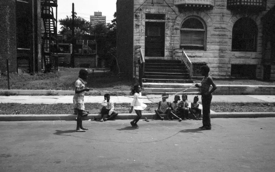 Four children play double dutch jump rope on a city street while five other children sit on the curb in front of a stone building with stairs and arched windows. It is a sunny day and the scene feels lively and communal.