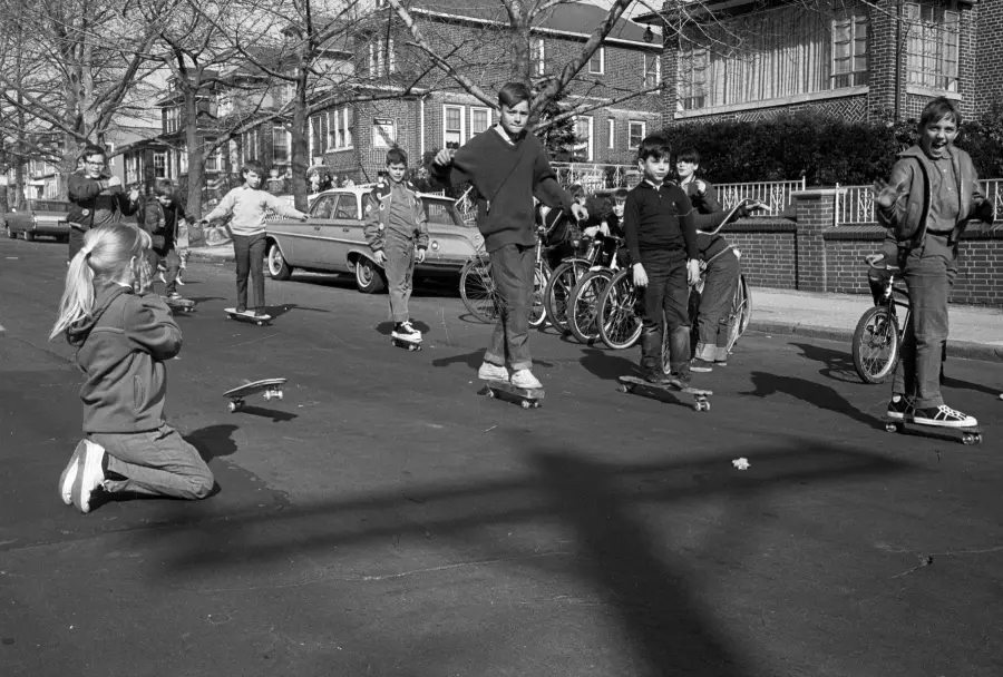 A group of kids ride skateboards and bikes on a suburban street in front of parked cars, with houses and trees in the background. One girl kneels on the ground, facing the group.