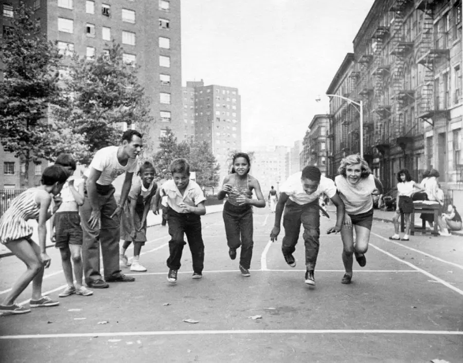 A group of children and two adults line up for a race on a city street, surrounded by tall apartment buildings and trees, on a sunny day.