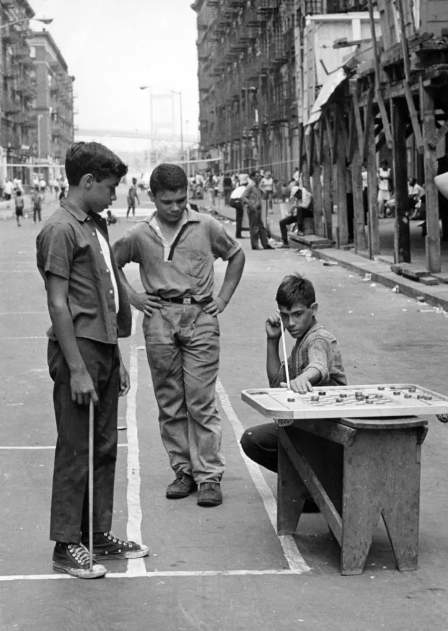 Three boys play a board game on a street table while others gather in the background; the scene appears to be from a mid-20th-century city neighborhood.