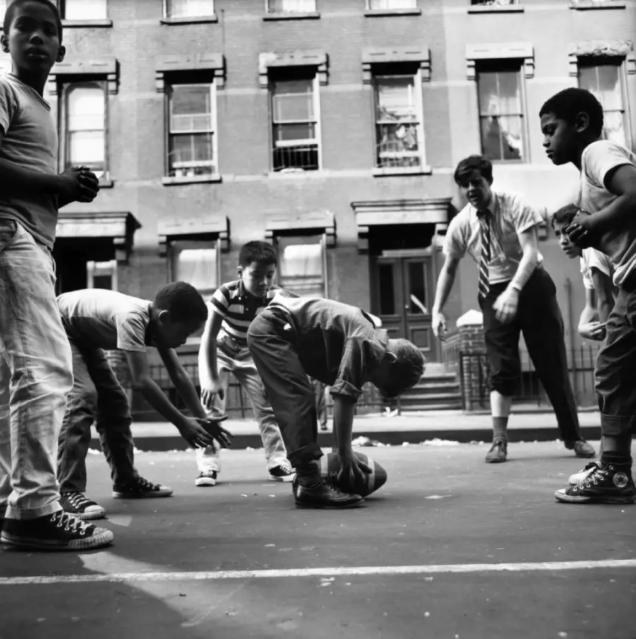 Black-and-white photo of children playing with a football on a city street, with an adult supervising. Brick apartment buildings line the background. The children appear energetic and engaged in the game.