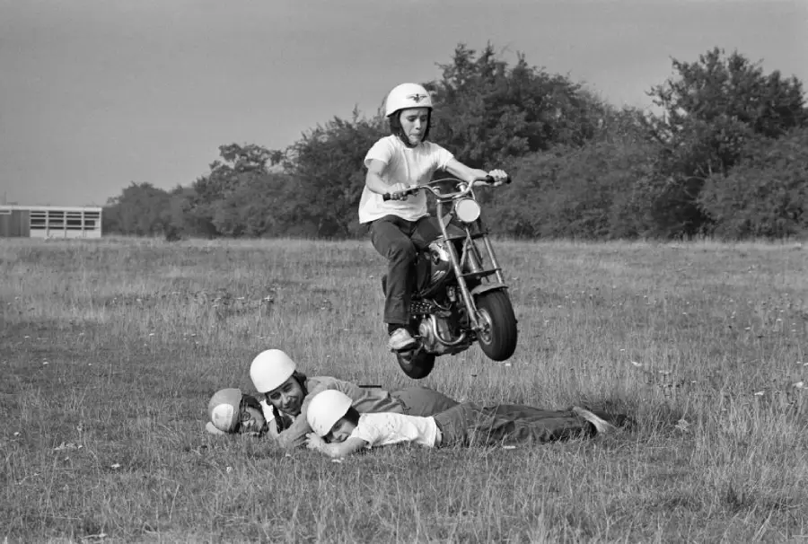 A person on a small motorcycle jumps over two people lying on the grass, all wearing helmets. The scene takes place outdoors in a grassy field with trees in the background.
