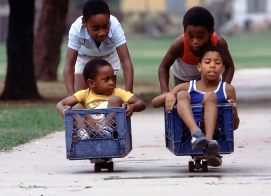 Four children play outside; two sit in milk crates on skateboards while the other two push them along a sidewalk. The scene is lively and joyful, set in a green park or neighborhood.