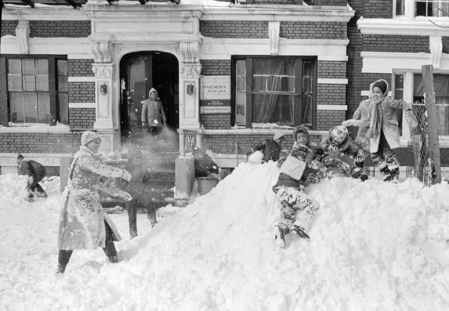 Children have a snowball fight and play on a large snow pile in front of an apartment building, while an adult stands in the doorway watching. Snow covers the ground and everyone appears to be having fun.