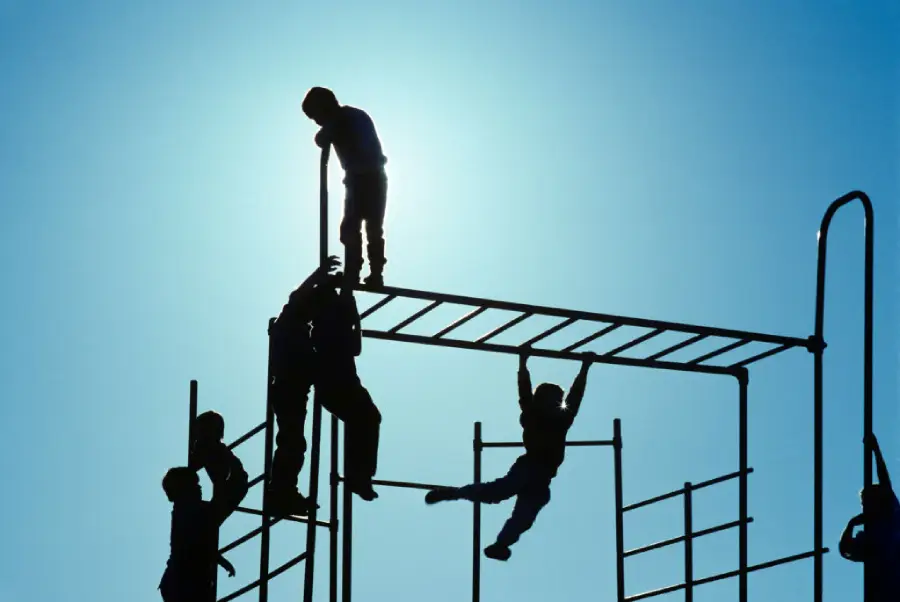 Silhouetted children climb and play on monkey bars at a playground against a bright blue sky, with the sun shining behind them.