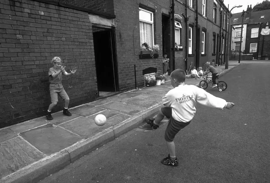 Two children play soccer on a sidewalk beside brick row houses, with one kicking the ball and the other ready to catch it. More kids ride tricycles further down the quiet street.