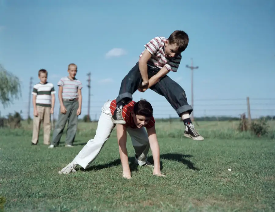 Four boys play leapfrog on a grassy field under a blue sky. One boy is jumping over another who is bent down, while two others watch in the background.