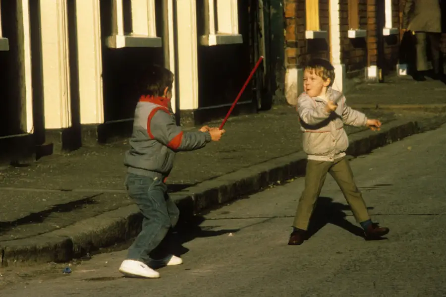 Two young boys play on a street; one swings a long red stick while the other dodges. Both wear jackets and pants. Buildings with black and white trim are visible in the background.