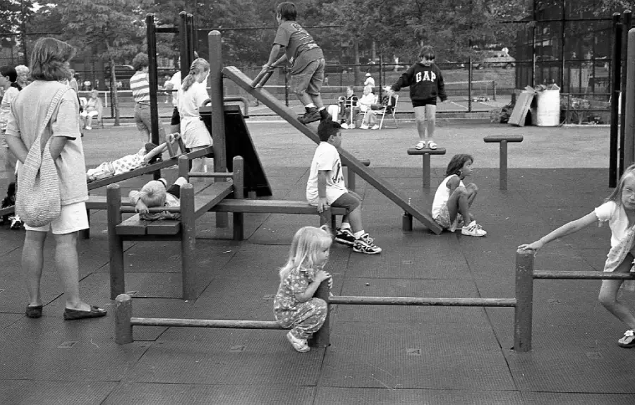 Children play on a playground with various equipment; some climb, others sit or balance. Adults watch nearby. The scene is outdoors with trees and fencing in the background.