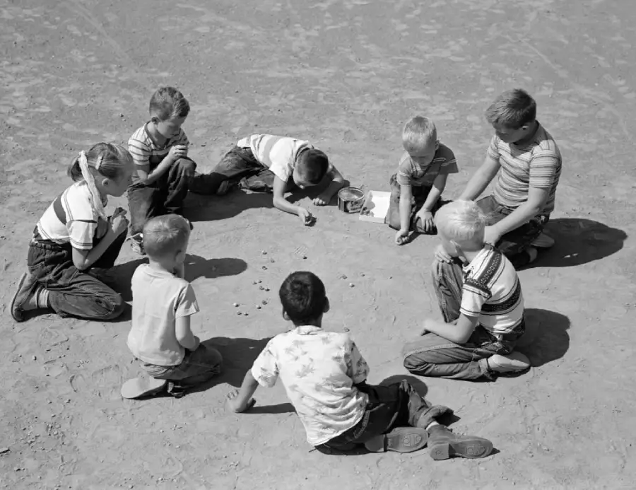 Seven children sit in a circle on dirt, intently playing a game with marbles. The scene appears to be outdoors on a sunny day, and the kids look focused and engaged.