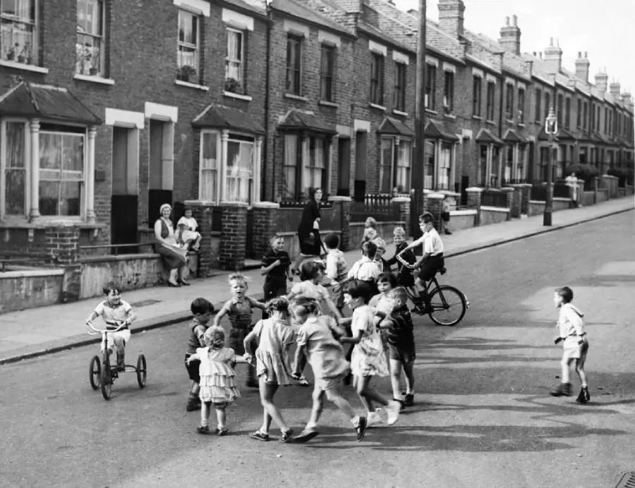 A group of children play and ride bikes on a residential street lined with terraced houses, while a few adults sit on steps and watch them. The scene appears to be from a past decade, giving a nostalgic atmosphere.